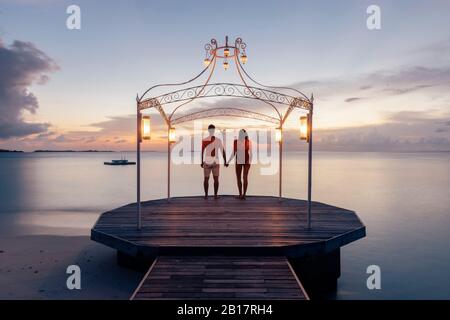 Couple standing on jetty at the sea at sunset, Maguhdhuvaa Island, Gaafu Dhaalu Atoll, Maldives Stock Photo