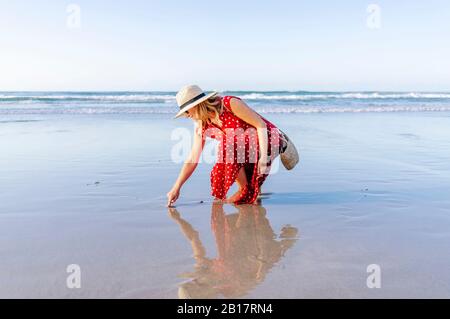 Blond woman wearing red dress and hat at the beach, Playa de Las Catedrales, Spain Stock Photo