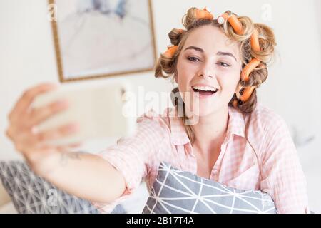 Portrait of happy of young woman with curlers in hair taking selfie with smartphone