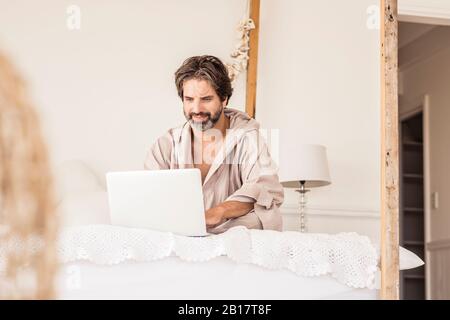Man wearing bathrobe and sitting on bed in bedroom Stock Photo