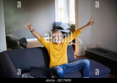 Mature man sitting on couch at home cheering Stock Photo
