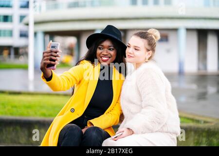 Happy girlfriends sitting on a bench in the city taking a selfie Stock Photo