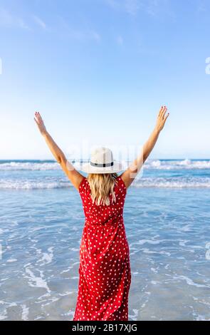 Blond woman wearing red dress and hat and walking along beach, Playa de Las Catedrales, Spain Stock Photo