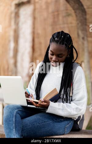 Female student using laptop and taking notes on a bench Stock Photo