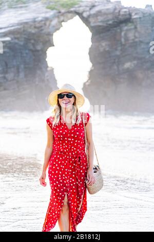 Blond woman wearing red dress and hat and walking along beach, Natural Arch at Playa de Las Catedrales, Spain Stock Photo