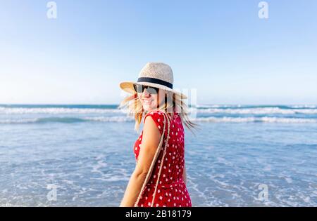 Blond woman wearing red dress and hat at the beach, turning and looking at camera Stock Photo