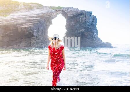 Blond woman wearing red dress and hat and walking along beach, Natural Arch at Playa de Las Catedrales, Spain Stock Photo