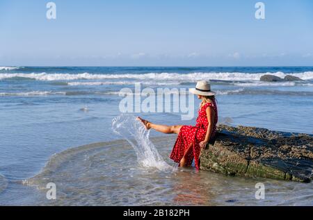 Blond woman wearing red dress and hat sittig on rock at the beach, Playa de Las Catedrales, Spain Stock Photo