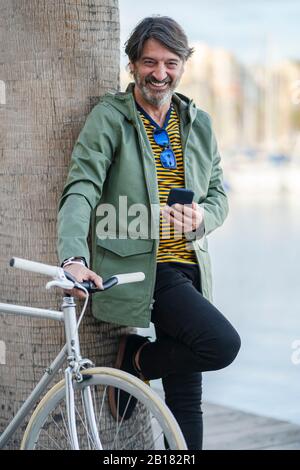 Portrait of laughing mature man with fixie bike leaning against palm tree trunk, Alicante, Spain Stock Photo