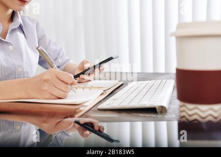 Young businesswoman reading messages in smartphone and taking notes in planner Stock Photo