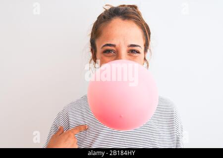 Young redhead woman chewing gum and blowing hair bubble over white isolated background with surprise face pointing finger to himself Stock Photo