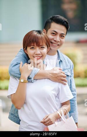 Happy young Asian man hugging his pretty girlfriend with many shopping bags from behind when that are standing outdoors Stock Photo