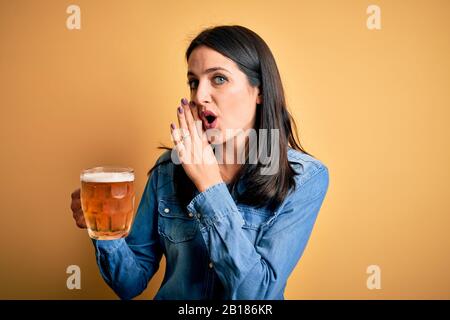 Young woman with blue eyes drinking jar of beer standing over isolated yellow background hand on mouth telling secret rumor, whispering malicious talk Stock Photo