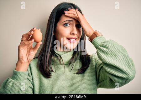 Young brunette woman with blue eyes holding fresh raw egg over isolated background stressed with hand on head, shocked with shame and surprise face, a Stock Photo