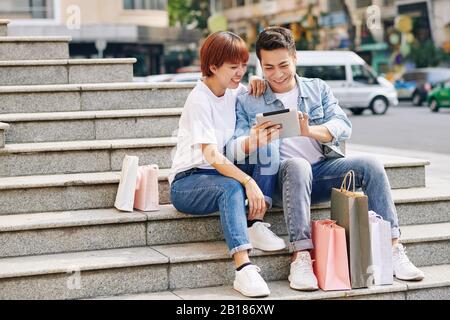 Cheerful young Vietnamese couple sitting on steps and making purchases via application on tablet computer Stock Photo