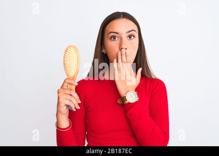 Young beautiful girl holding hair comb standing over isolated white background cover mouth with hand shocked with shame for mistake, expression of fea Stock Photo