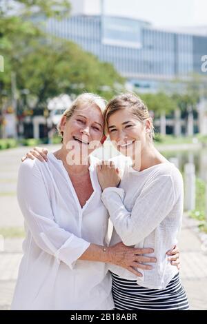 Happy mature woman and her adult daughter hugging and looking at camera when standing in city park Stock Photo