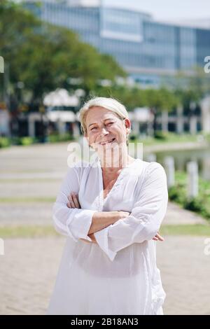 Portrait of pretty smiling mature woman standing at city park with arms folded and looking at camera Stock Photo
