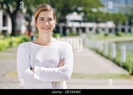 Lovely mixed-race young woman in white sweater standing in city park with arms folded and looking at camera Stock Photo