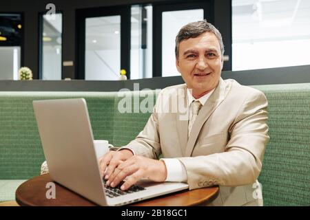 Portrait of positive middle-aged businessman working on laptop and smiling at camera Stock Photo