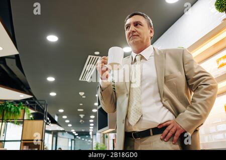 Serious pensive mature entrepreneur standing in office with big mug when having coffee break Stock Photo