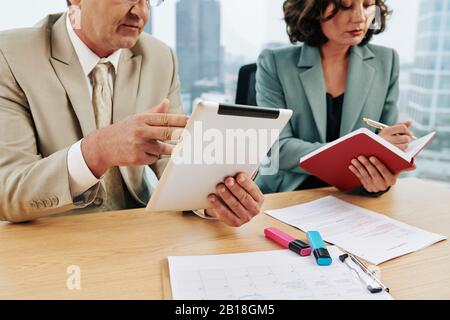 Businessman reading report on talet computer when his colleague writing down important thoughts in planner Stock Photo