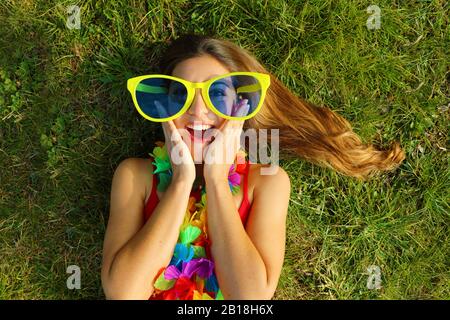 Surprised girl wake up on grass after Carnival party. Happy excited oung woman with carnival garland and big funny sunglasses lying on grass. Stock Photo