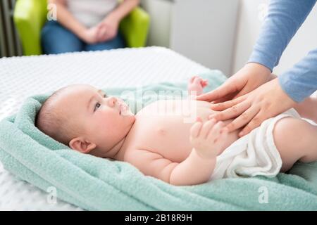 Female massage therapist or a doctor examining newborn baby boy with the mother watching in the background. Baby massage concept. Stock Photo