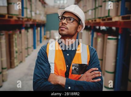 Portrait of serious supervisor in warehouse standing with folded hands looking high at shelves with material while wearing orange vest and white Stock Photo