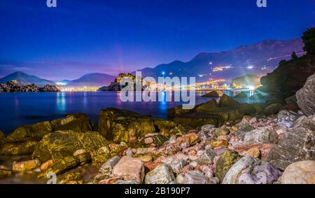 Stones on the coast of Adriatic sea near illuminated Sveti Stefan at sunset, Montenegro Stock Photo