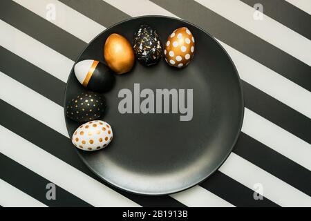 Black and white striped background with white, black and golden decorated eggs on black plate. Trendy easter flat lay. Stock Photo