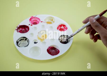 A person taking some paint with a brush from a color pallet to paint something ,over a yellow paper background Stock Photo