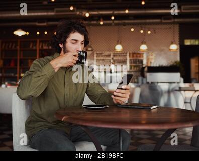 Handsome young caucasian man browsing through smartphone while drinking coffee in cafeteria Stock Photo
