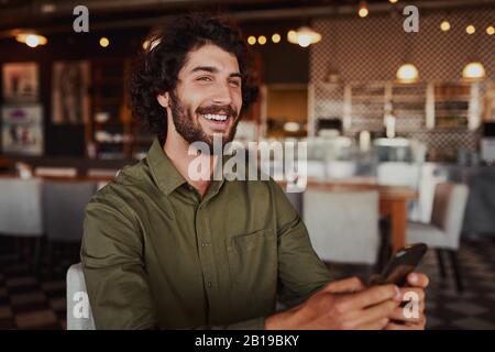 Portrait of handsome young man laughing while reading funny message on phone sitting in modern cafeteria during free time Stock Photo