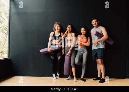 Happy smiling man and women looking at camera altogether in gym. Group of young people relaxing in gym after workout training with black background. Stock Photo