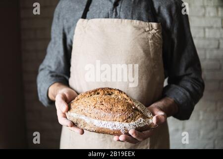 Man in rustic apron holding a loaf of bread in his hands. Close up on the product. Stock Photo