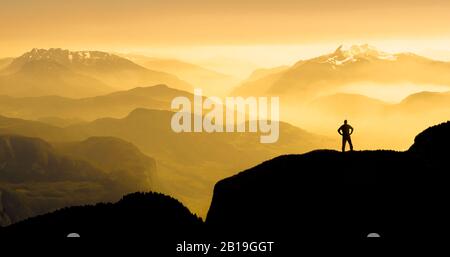 Spectacular mountain ranges silhouettes. Man reaching summit enjoying freedom. Sunrise with orange light. Stock Photo