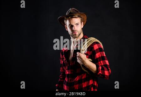 Western life. Lassoing on prairie. Man unshaven cowboy black background. Cowboy wearing hat hold rope. Lasso tool of American cowboy. Still used on ranches to capture cattle or other livestock. Stock Photo