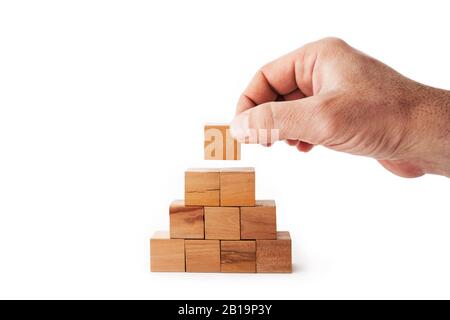 A hand is putting the last cube for building a pyramid with wooden cubes on white background. Success, strategy, improvement and victory concept Stock Photo