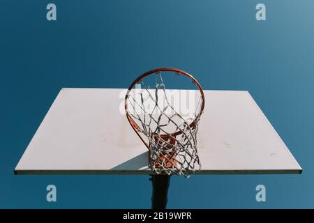 Looking up at old basketball hoop with torn net and white backboard alone against blue sky. Stock Photo
