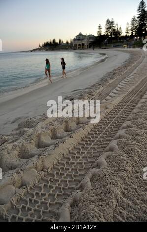 Tire tracks in sand, with two young women walking in shallows, Cottesloe Beach, Perth, Western Australia Stock Photo