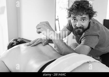 Black and white image of a curly-haired male physiotherapist doing therapeutic body massage to a strong male patient. Desaturated picture Stock Photo