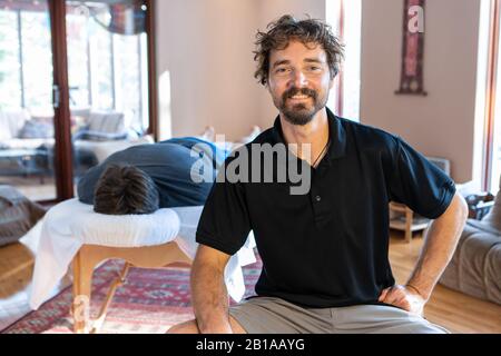 Portrait of smiling physiotherapist on working place. A strong male client lying on the table and waiting for treating body massage Stock Photo