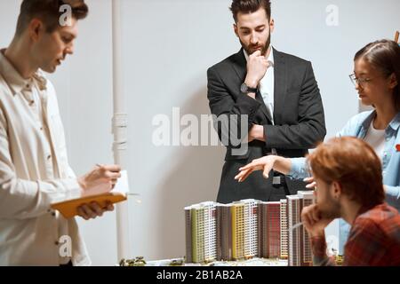 clever woman in glasses gesturing, talking her colleagues about creative design and construction of building, bearded man touching his chin, listening Stock Photo