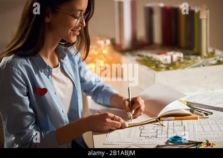 business woman gets plasure from architectural work, pretty woman making notes while working on a project. close up side eview photo. small model of d Stock Photo