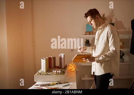 young architect holding yellow book and glasses standing in the office, small layout in the background of the photo, close up side view photo Stock Photo
