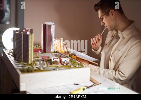 young pensive thoughtful designer with a pen in his mouth reading something from his notepad, guy biting his pen and holding a notebook, thinking abou Stock Photo