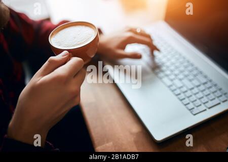 Man in cafe using laptop to chat and play social media and drinking hot coffee.close up cropped side view photo. male hands with coffee cup , man sitt Stock Photo