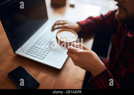 man's arms holding cup of coffee and typing on laptop. close up cropped photo, focus on a cup with foam, art Stock Photo