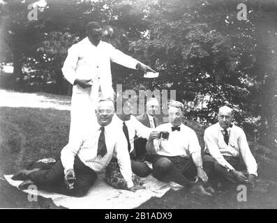 One African American waiter/ server holds up a cash tip he received from 5 White businessmen sitting on a picnic blanket. My guess is that the Whites told him to display the cash for the photographer, to show how generous they were. I can't see what amount the cash tip is. The businessmen are enjoying beer, having a good time.  No known location or people.   To see my other African American-related vintage images, Search:  Prestor  vintage  African Stock Photo